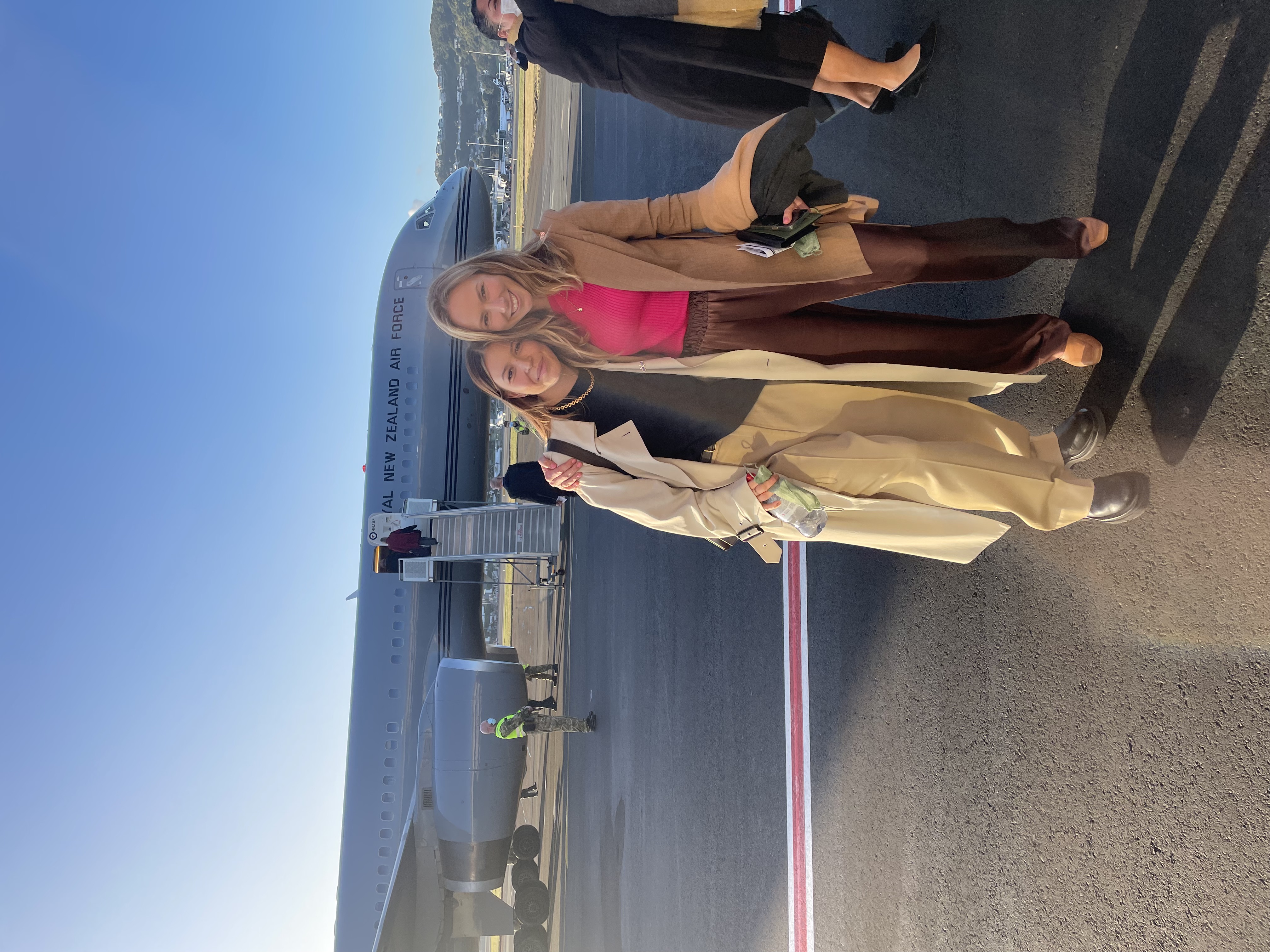 Brooke Roberts and Jessie Wong are smiling into the camera, standing on an airport runway in front of a Royal New Zealand Air Force plane. 