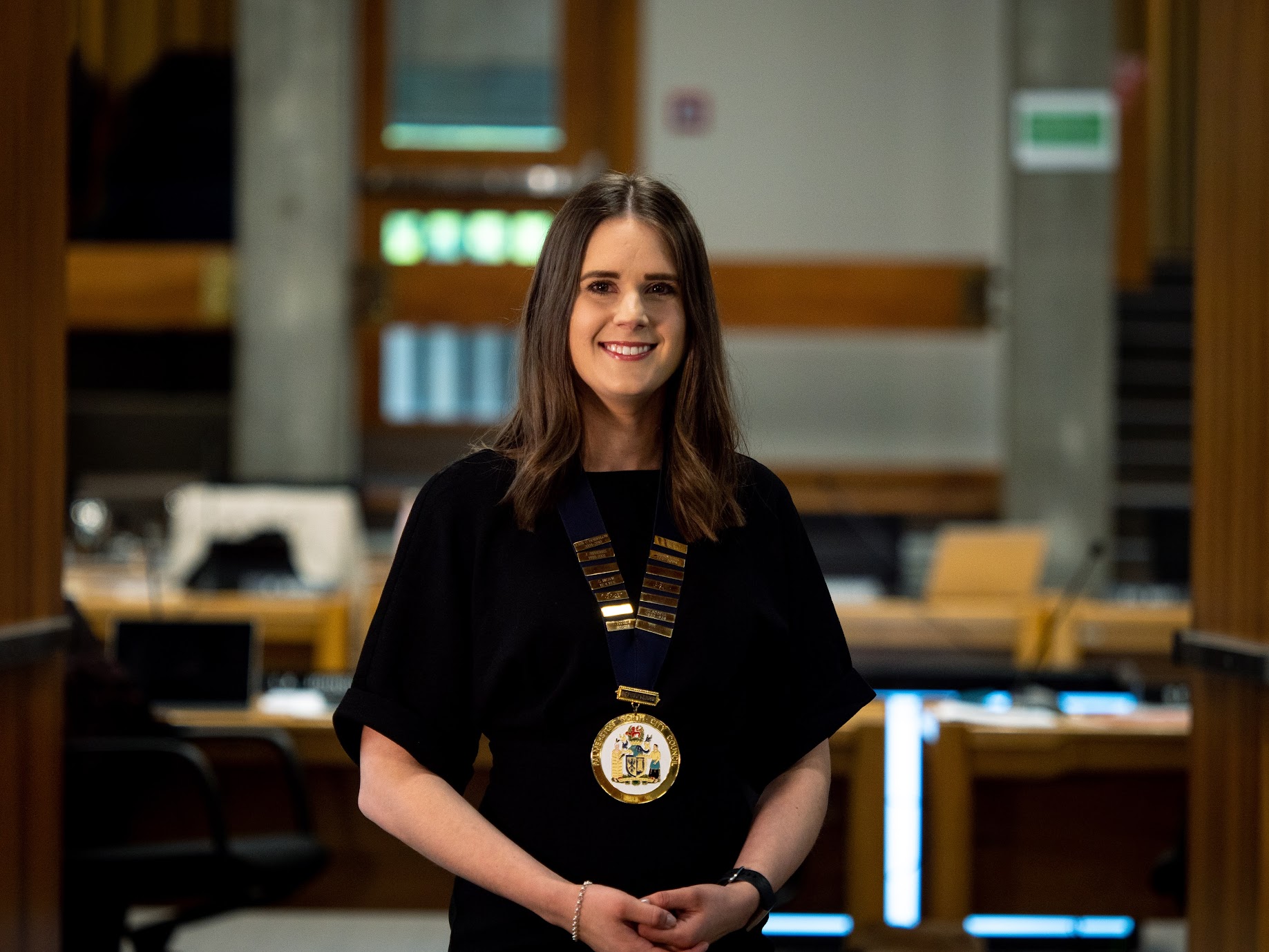 Aleisha is wearing her mayoral chain, and smiles toward the camera.