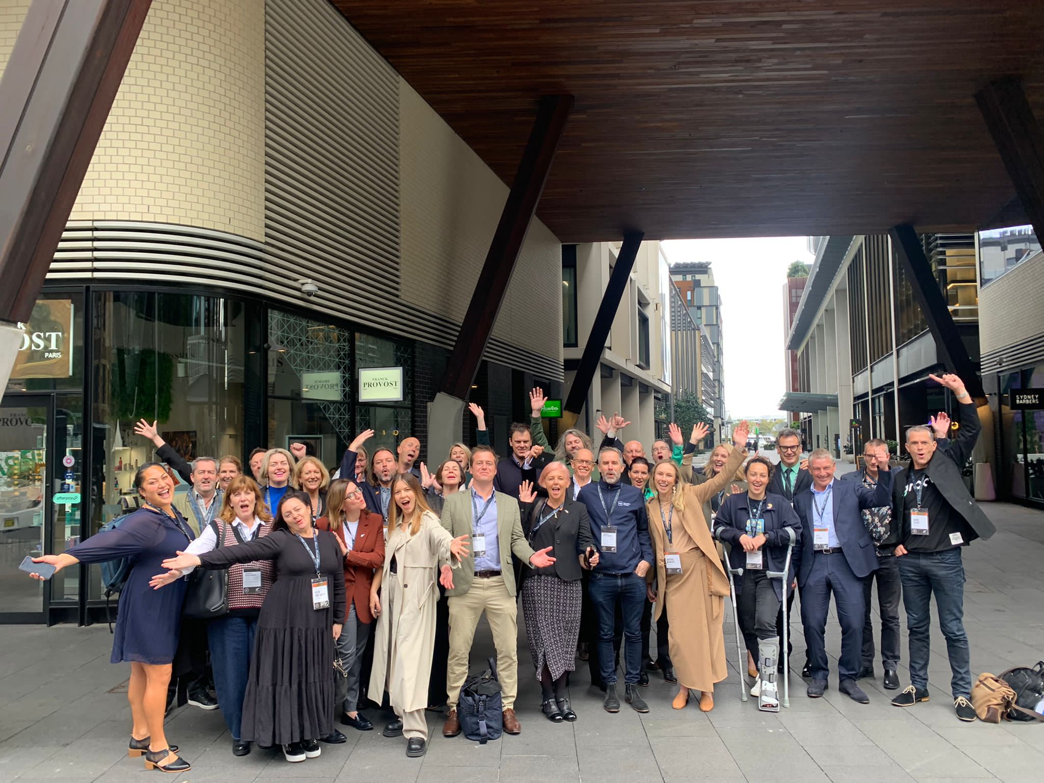 The full trade delegation standing outside of a city building, all smiling and looking into the camera with their arms wide open. 