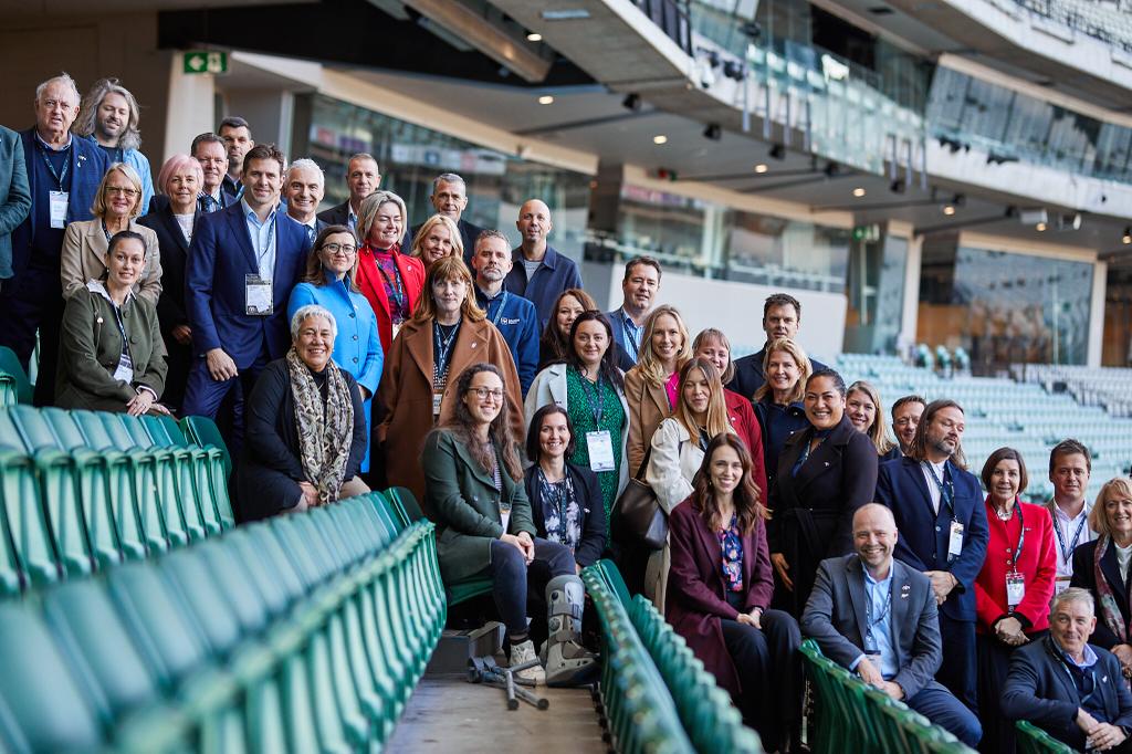A picture of the full trade delegation, standing in a row of stadium seating. Everyone is standing from the top left of the photo to the bottom right, all looking into the camera and smiling. 