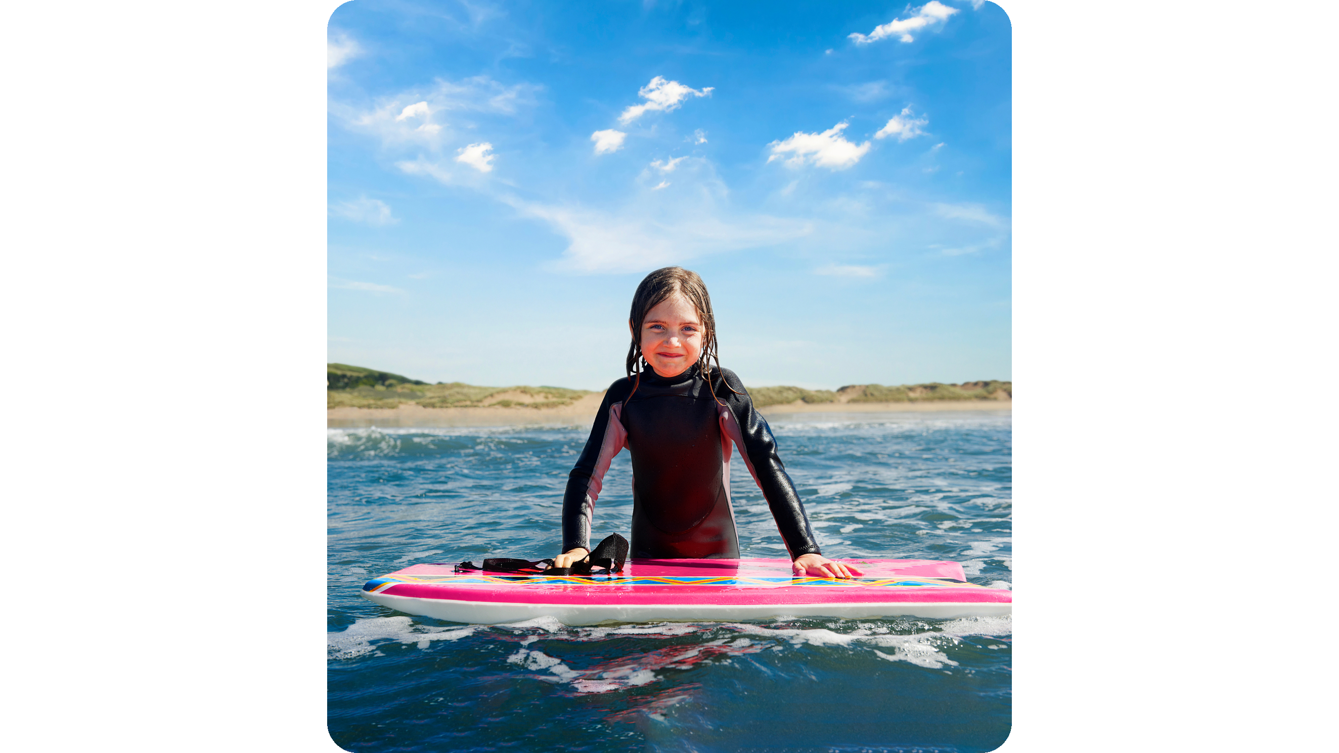 A young girl in a wet suit stands in the sea with her hands on a boogie board.